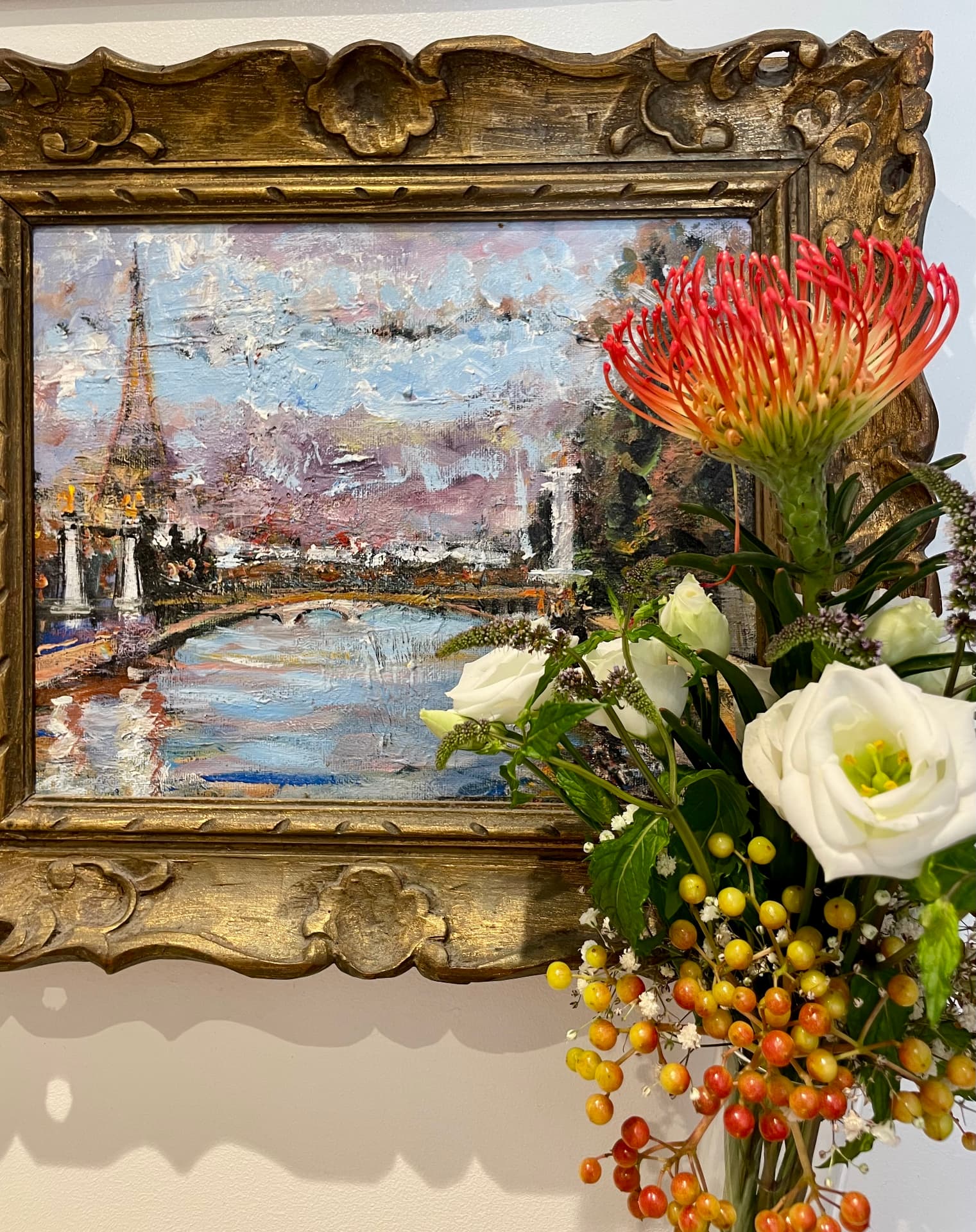 Gilded frame with an oil painting of the Eiffel Tower beside protea, white roses and berries