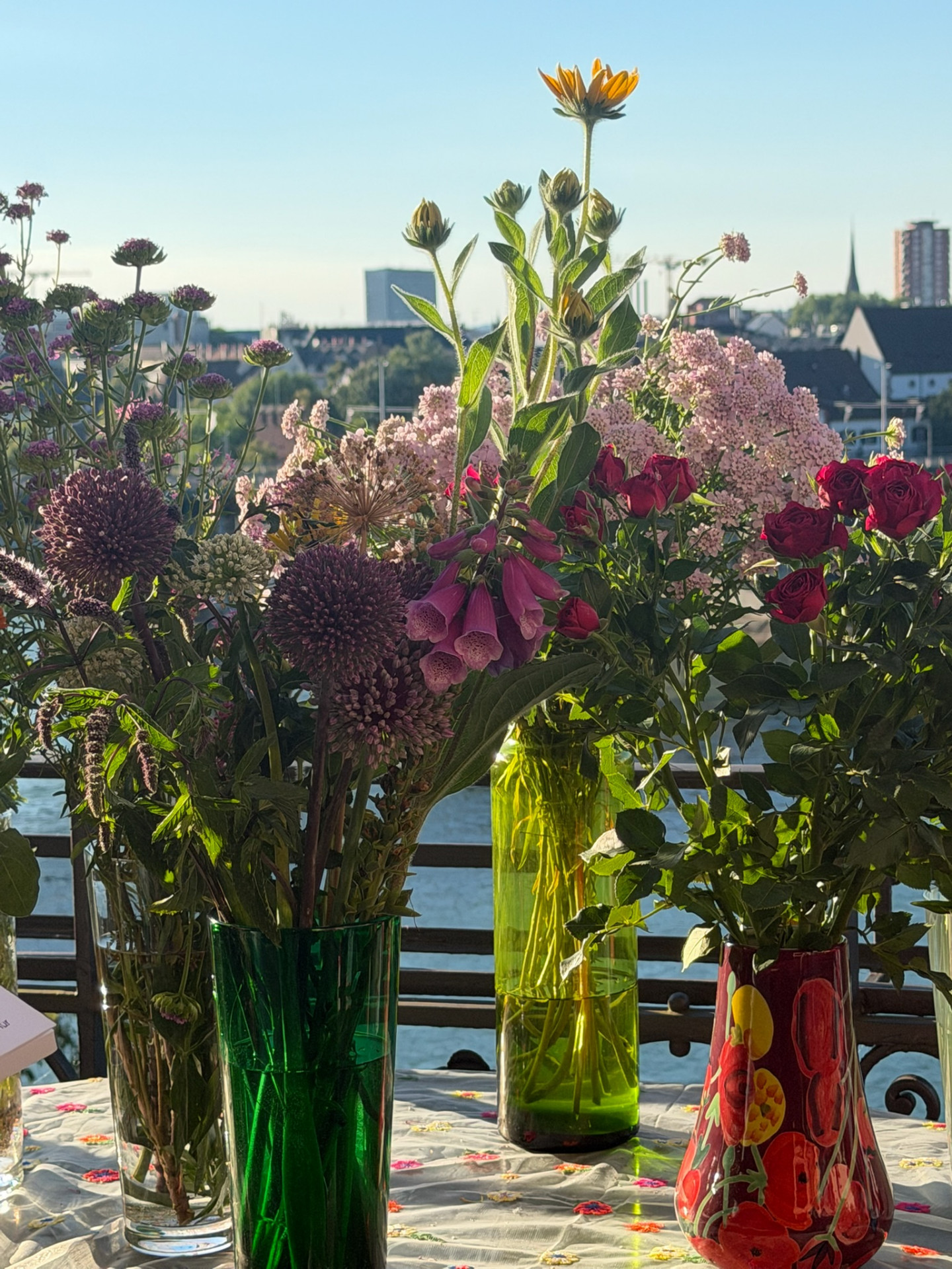 Close-up of allium, foxglove, sunflower and red roses in coloured glass vases against the Basel skyline