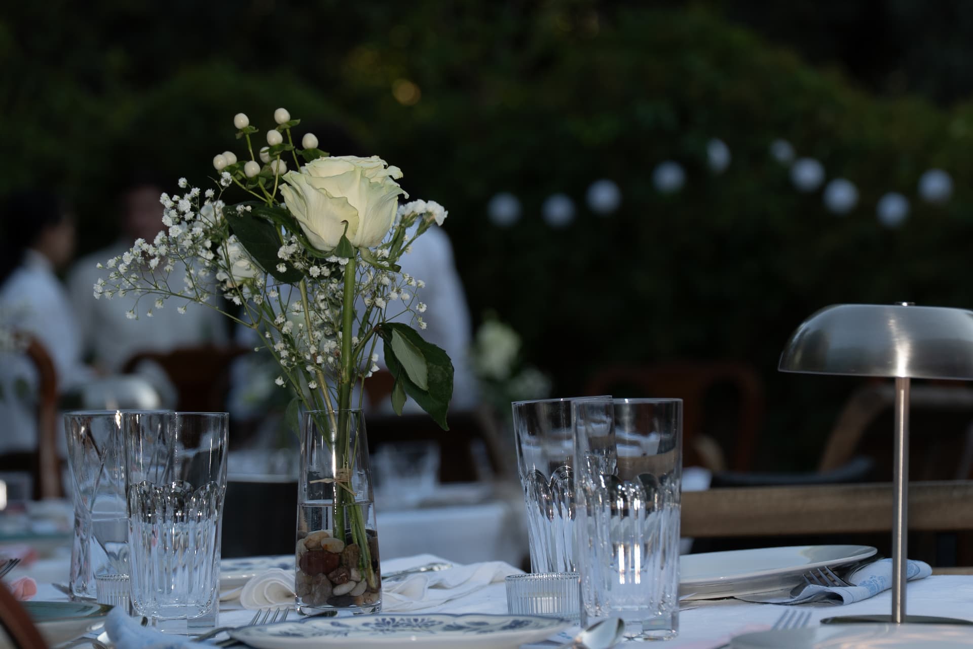 White rose and gypsophila arrangement on the candlelit table with festoon lights in the garden behind