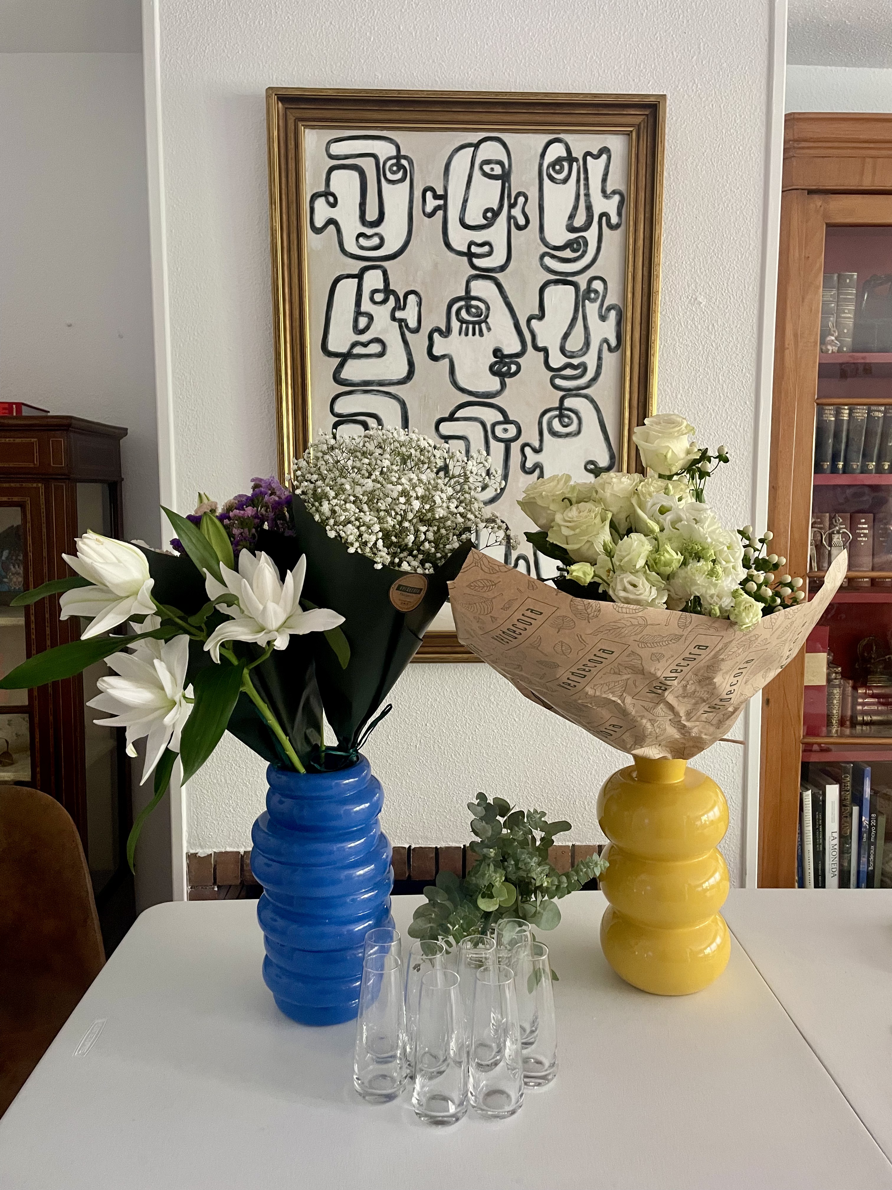 Two wrapped bouquets of white lilies, gypsophila, roses and lisianthus in blue and yellow ribbed vases beneath a line-drawn portrait painting