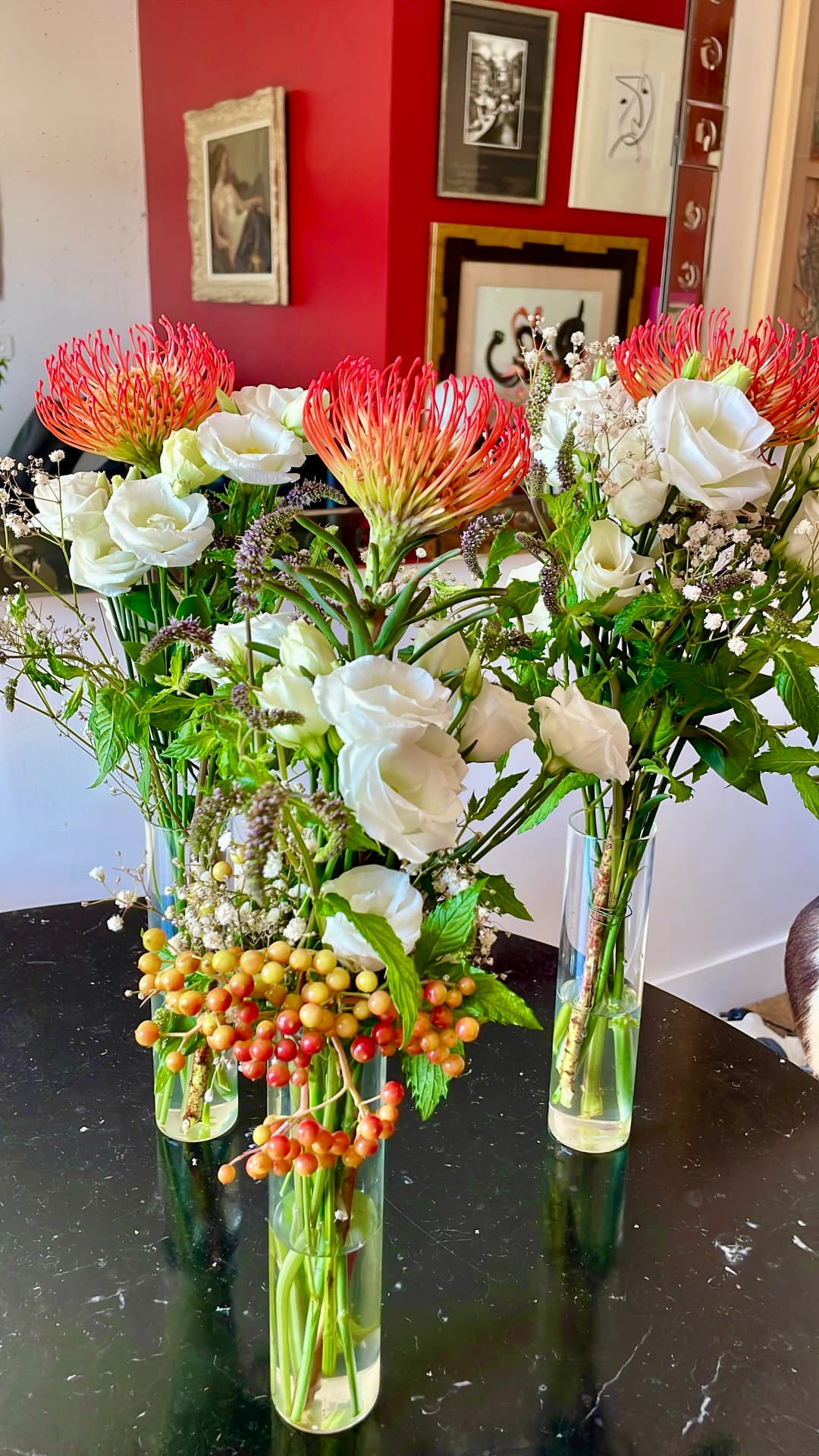 Three glass bud vases of pincushion protea, white lisianthus, mint and berries on a black marble table