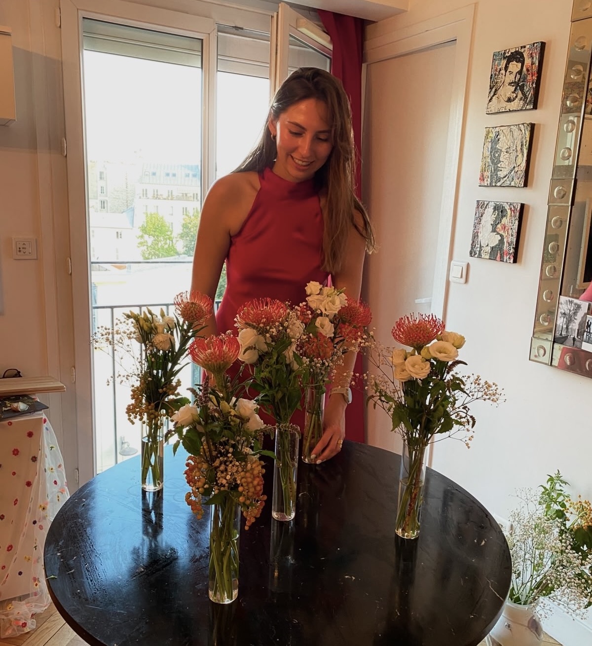 Host in a red dress smiling beside bud vases of pincushion protea and white lisianthus on a black table in a Paris apartment
