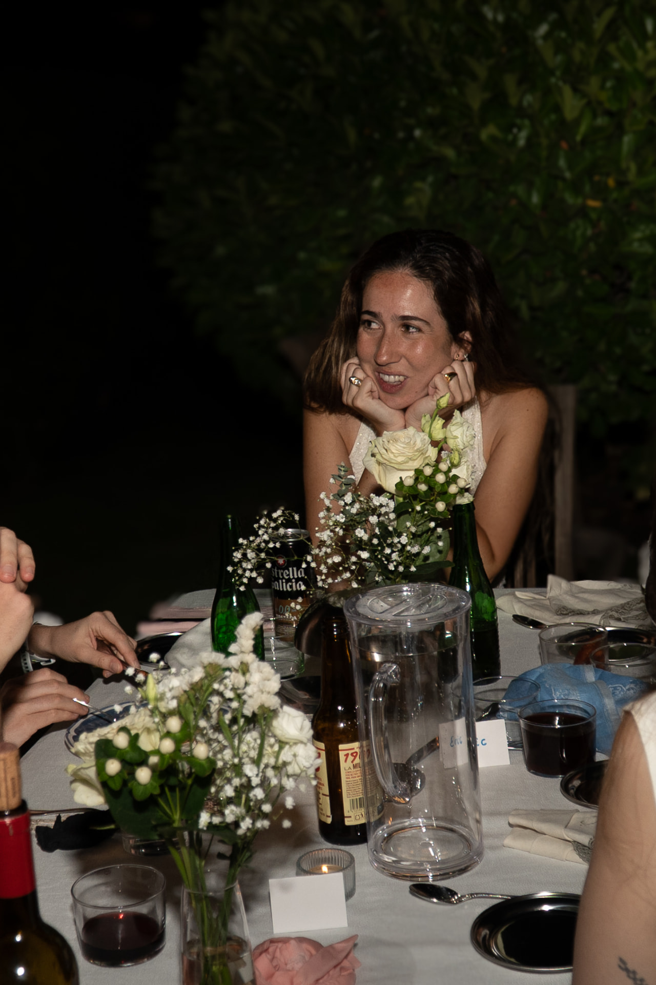 Guest resting her chin on her hands behind bud vases of white roses at the night-time dinner table