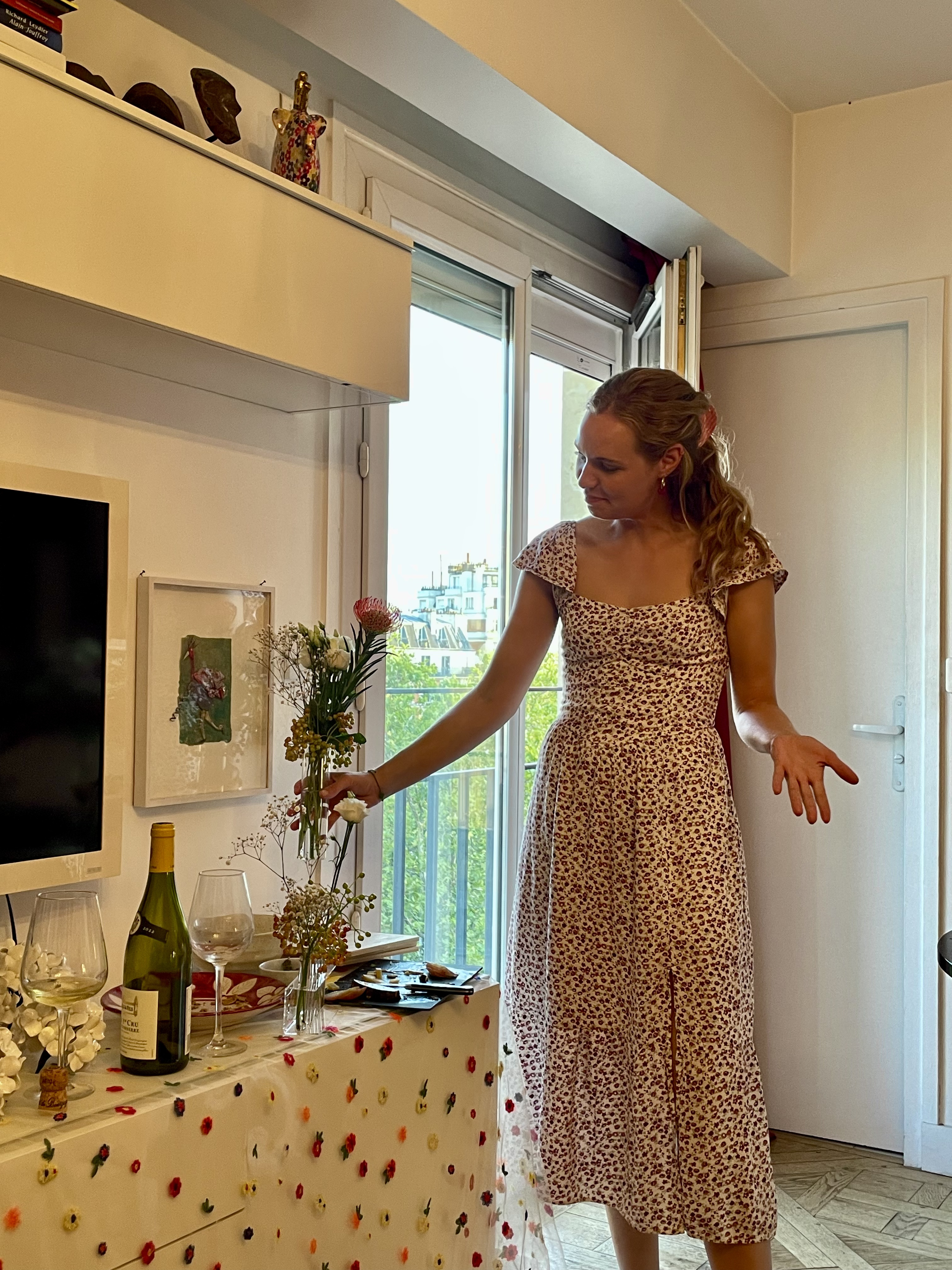 Host in a floral dress gesturing towards bud vases of pincushion protea and white roses by a Paris window