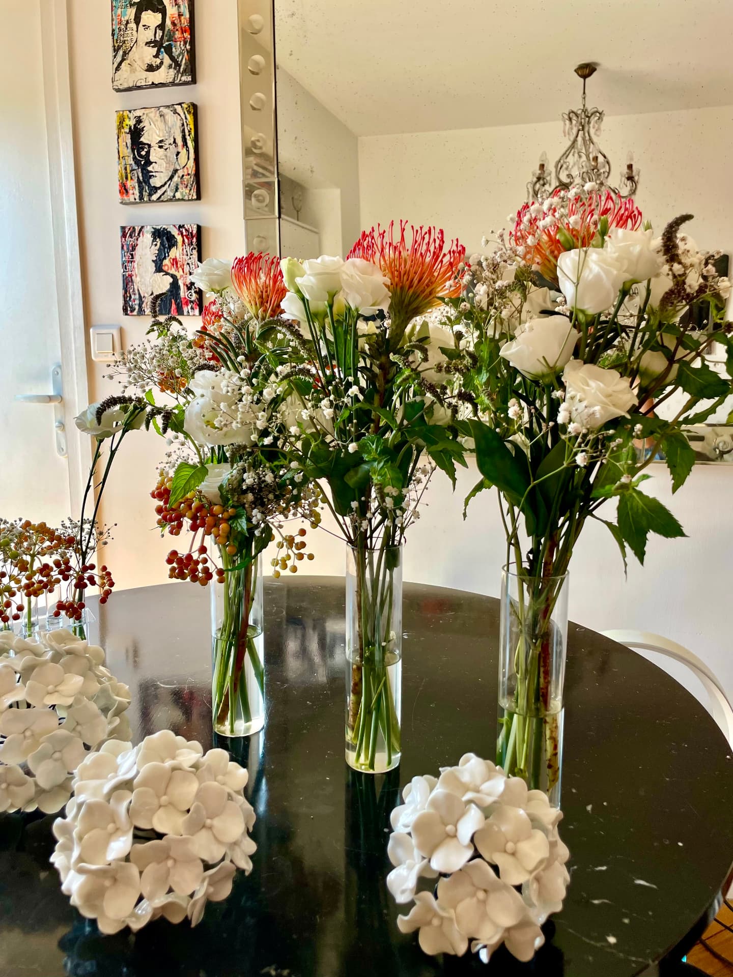Three tall arrangements of protea, white roses and gypsophila on a round black table beside ceramic hydrangeas