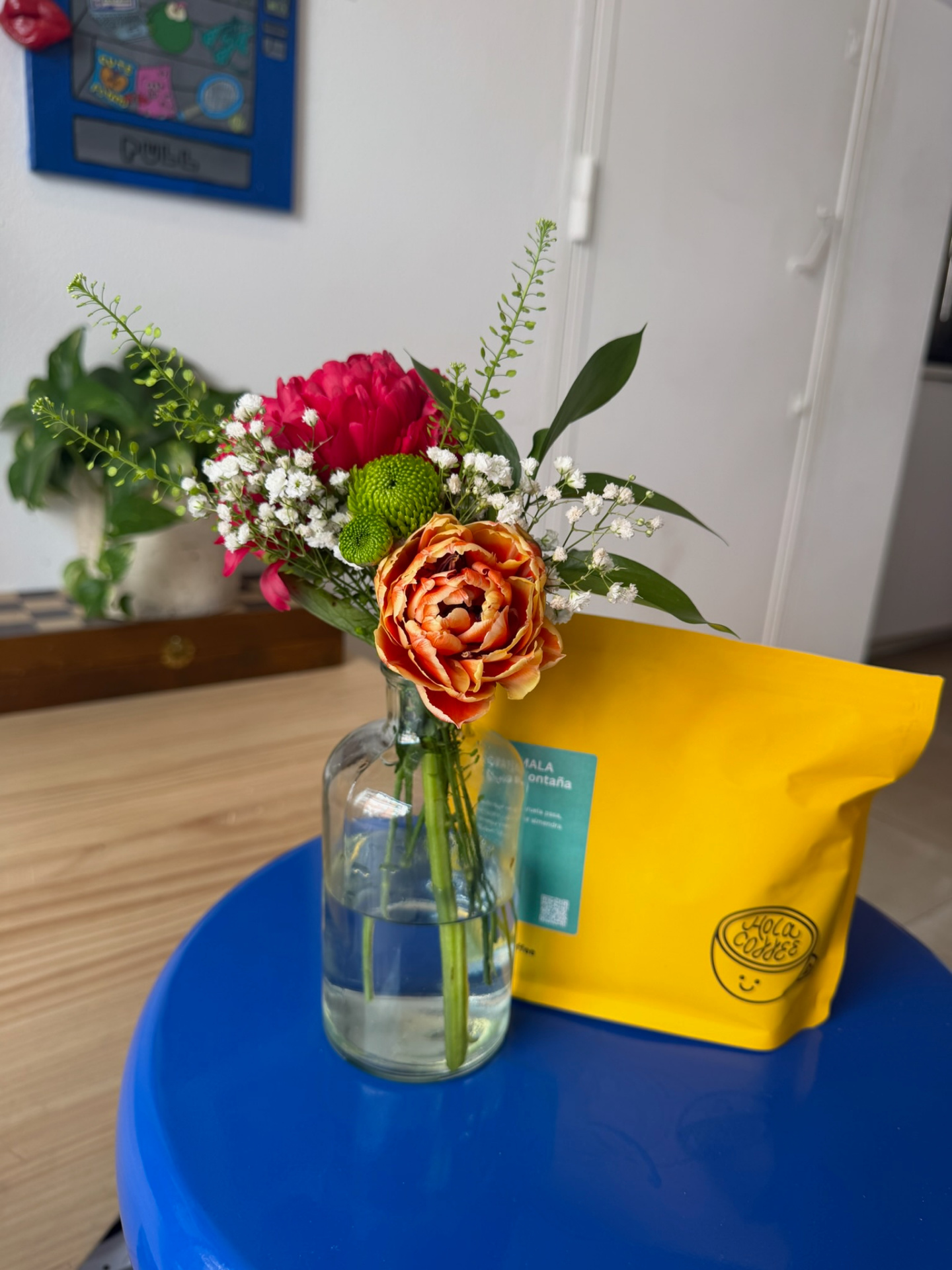 Bud vase with orange tulip, pink chrysanthemum and greens beside a yellow coffee bag